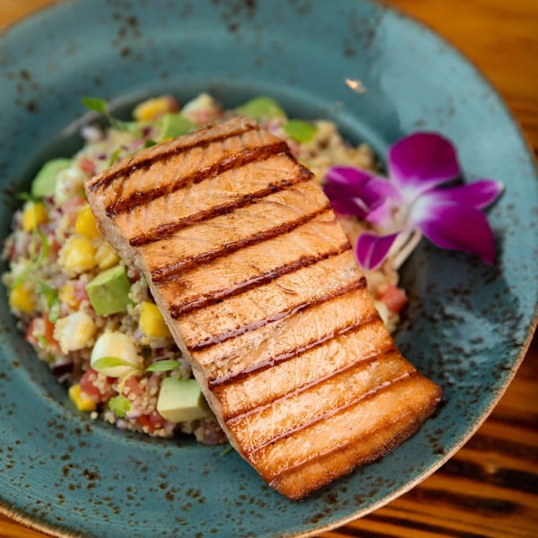 Grilled salmon with quinoa salad, avocado, corn, and a purple orchid garnish on a blue plate.