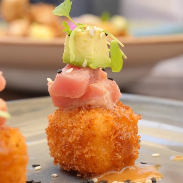 Close-up of a golden-fried Peruvian causa bite topped with tuna, avocado, and sesame seeds.