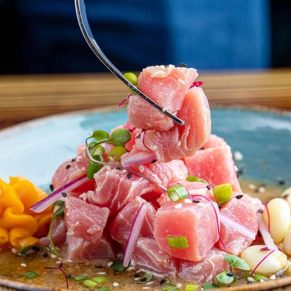 Fork lifting a piece of tuna ceviche from a blue plate with red onions and corn.